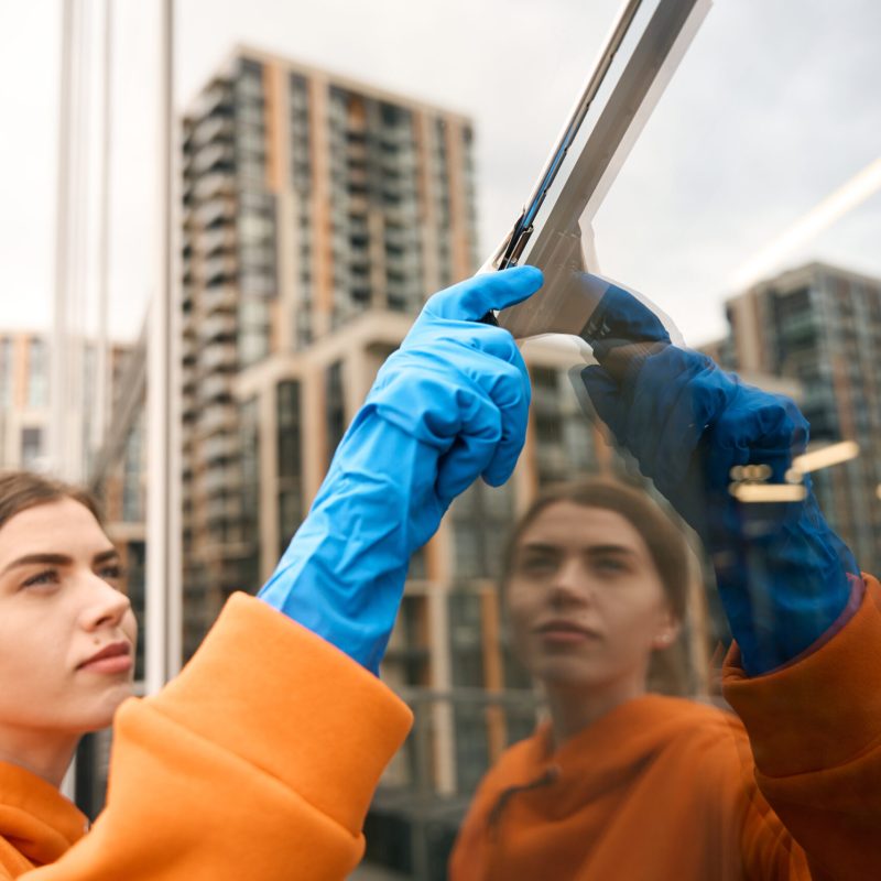 Cleaning service cleaner cleaning the mirrored windows of the office center with a glass scraper, she is wearing rubber gloves