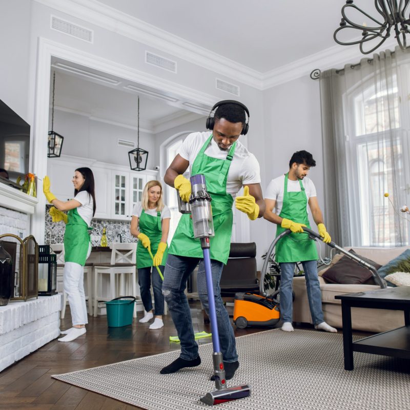 Handsome young african worker of cleaning service, cleaning up carpet and dancing, listening to music in headphones, while another members of cleaning team working on background.