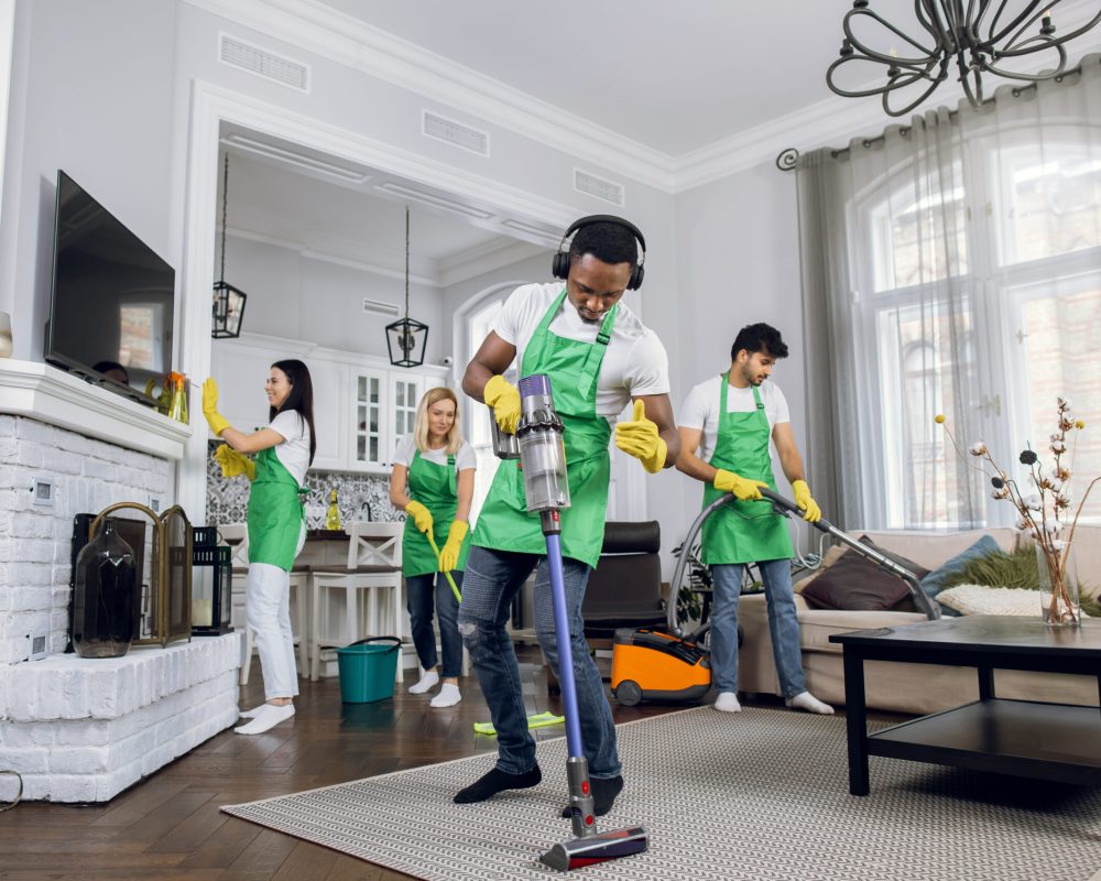 Handsome young african worker of cleaning service, cleaning up carpet and dancing, listening to music in headphones, while another members of cleaning team working on background.