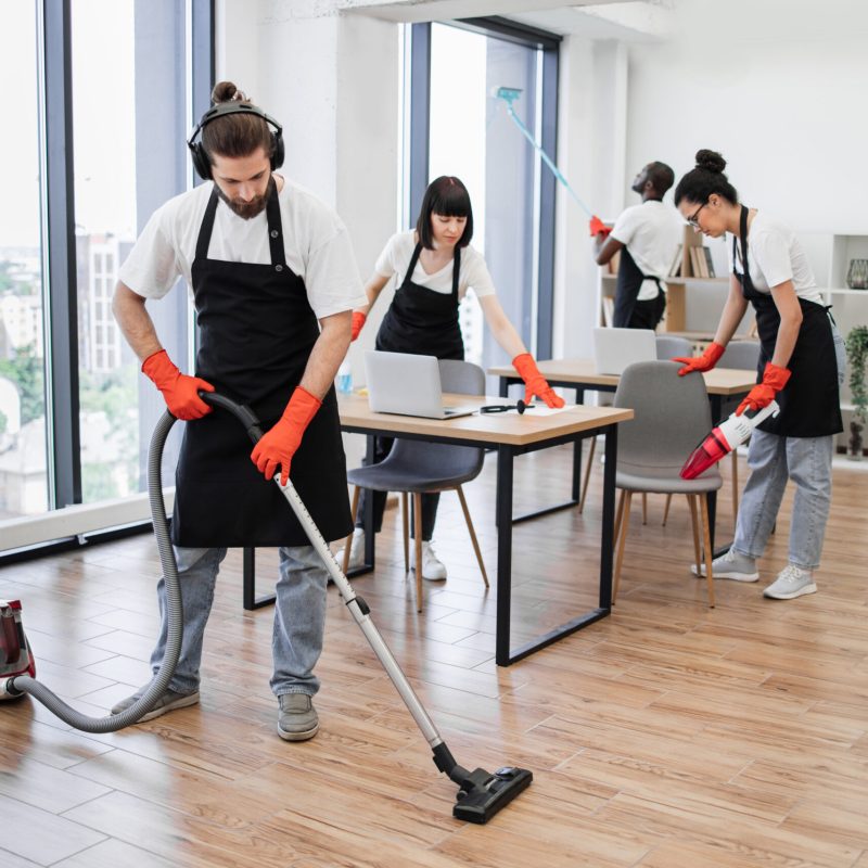 Team of young multicultural cleaners vacuums floor, wipes tables with gadgets, shelves, washes windows in spacious, bright, modern office. Professional cleaning company.