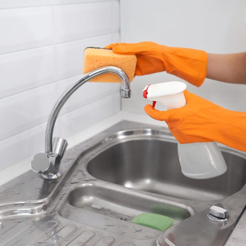 Close up of woman in orange rubber gloves cleaning kitchen faucet. Concept of cleaning in kitchen.